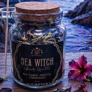 May include: A clear glass jar with a cork lid containing loose tea, labeled Sea Witch Specialty Loose Tea, Pea Flower, Hibiscus, Lemongrass, 8 oz. A thin wooden stick rests against the jar, surrounded by scattered dried tea ingredients.