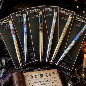 May include: Six ballpoint pens with clear barrels filled with colourful crystals, each mounted on a black display card labelled with names like Bliss, Guardian, Serenity, Prosperity, and Empower, arranged behind an open book with ritual text.