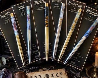 May include: Six ballpoint pens with clear barrels filled with colourful crystals, each mounted on a black display card labelled with names like Bliss, Guardian, Serenity, Prosperity, and Empower, arranged behind an open book with ritual text.