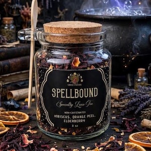 May include: A clear glass jar with a cork lid containing loose leaf tea. The black label reads SPELLBOUND Specialty Loose Tea, NON-CAFFEINATED, HIBISCUS, ORANGE PEEL, ELDERBERRY, 8 oz. A wooden spoon rests in the jar handle. Dried tea leaves are visible.