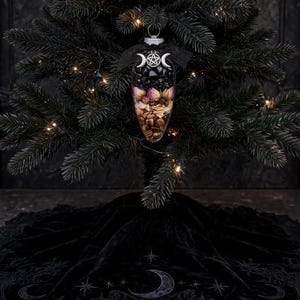 May include: A clear, teardrop-shaped glass ornament filled with black stones, dried pink rosebuds, and brown tiger eye crystals. The top features a silver pentacle charm flanked by two crescent moons, finished with a black ribbon bow.