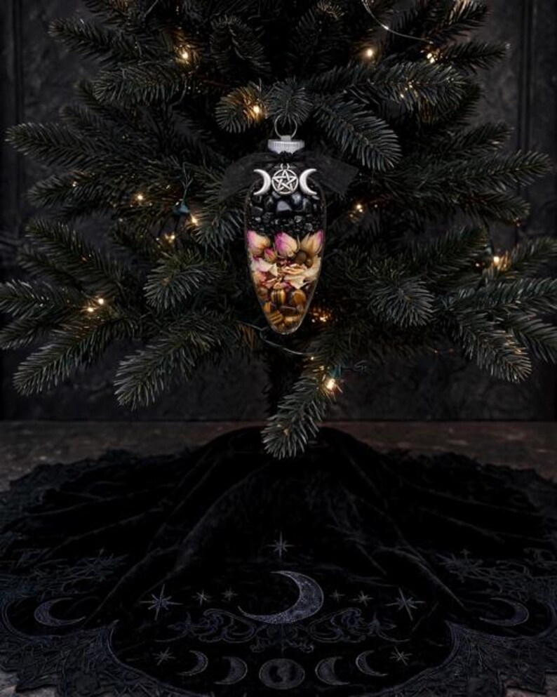 May include: A clear, teardrop-shaped glass ornament filled with black stones, dried pink rosebuds, and brown tiger eye crystals. The top features a silver pentacle charm flanked by two crescent moons, finished with a black ribbon bow.