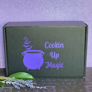 May include: A black box with the text "Cookin Up Magic" in white lettering and a purple cauldron with bats flying around it. The box is sitting on a black and white patterned surface.