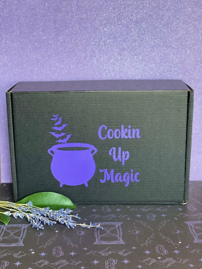 May include: A black box with the text "Cookin Up Magic" in white lettering and a purple cauldron with bats flying around it. The box is sitting on a black and white patterned surface.
