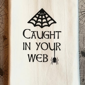 Op de afbeelding: Een witte keukendoek met zwarte tekst die "Caught in your web" luidt en een zwarte spinnenweb- en spinafbeelding.