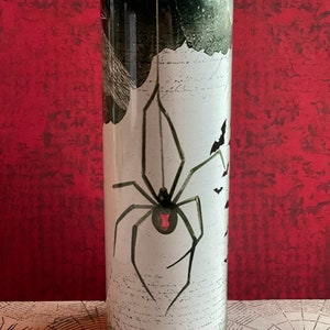 Op de afbeelding: Een zwarte en groene roestvrijstalen tumbler met een wit spinnen- en vleermuisontwerp. De spin heeft een rode buik en hangt aan een web. De achtergrond is een wit en zwart patroon.