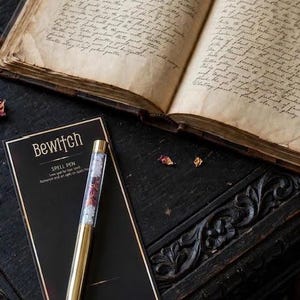 May include: A gold-toned ballpoint pen with a clear barrel filled with small crystals and dried petals, resting on a black display card labelled "Bewitch Spell Pen" with text listing Rose Quartz, Rose Petals, and Moonstone, beside an open antique book.