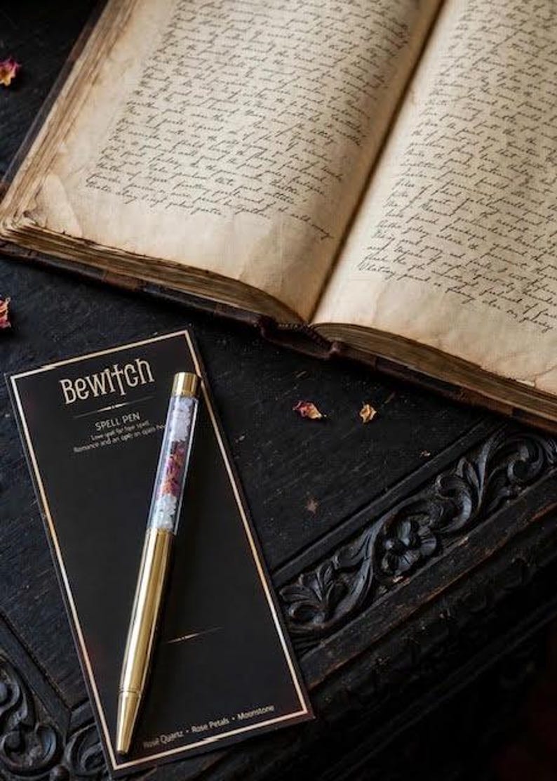 May include: A gold-toned ballpoint pen with a clear barrel filled with small crystals and dried petals, resting on a black display card labelled "Bewitch Spell Pen" with text listing Rose Quartz, Rose Petals, and Moonstone, beside an open antique book.