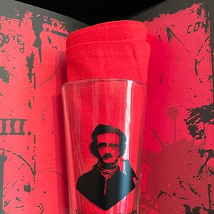 May include: A clear glass with a black silhouette of Edgar Allan Poe and a raven, with the text "Quoth the Raven Nevermore". The glass is sitting on a red and black patterned surface.
