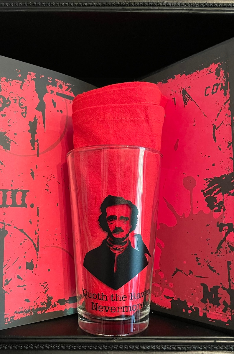 May include: A clear glass with a black silhouette of Edgar Allan Poe and a raven, with the text "Quoth the Raven Nevermore". The glass is sitting on a red and black patterned surface.