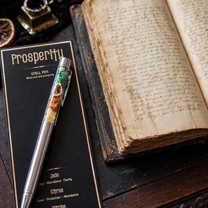 May include: A silver-toned ballpoint pen with a clear barrel filled with green jade stones, dried citrus slices, and yellow citrine crystals. The pen rests on a black card labelled Prosperity, Spell Pen, Jade, Citrus, and Citrine with descriptive text.