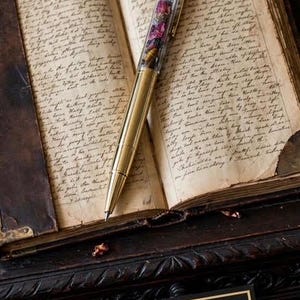 May include: A gold-toned metal pen with a clear barrel filled with small dark and purple stone fragments, resting on an open book with handwritten cursive text. A black rectangular card with the gold text Guardian Spell Pen is visible below.