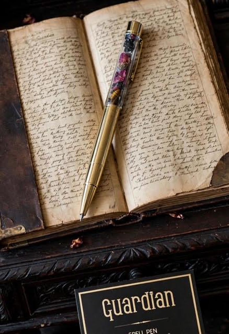 May include: A gold-toned metal pen with a clear barrel filled with small dark and purple stone fragments, resting on an open book with handwritten cursive text. A black rectangular card with the gold text Guardian Spell Pen is visible below.