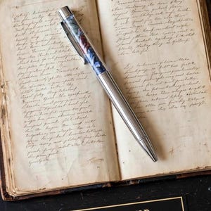 May include: A silver ballpoint pen with a clear barrel filled with small, polished blue and red stones. The pen features a metallic clip and a pointed tip, resting diagonally across an open book with handwritten cursive text on aged paper.