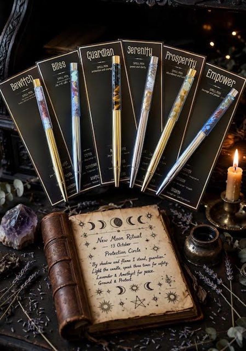 May include: Six ballpoint pens with clear barrels filled with colourful crystals, each mounted on a black display card labelled with names like Bliss, Guardian, Serenity, Prosperity, and Empower, arranged behind an open book with ritual text.