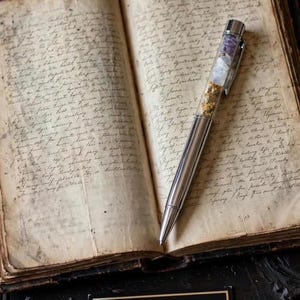 May include: A silver-toned ballpoint pen with a clear upper barrel containing small purple, white, and gold-coloured decorative fragments, resting on an open antique book with handwritten cursive text on aged, yellowed paper pages.