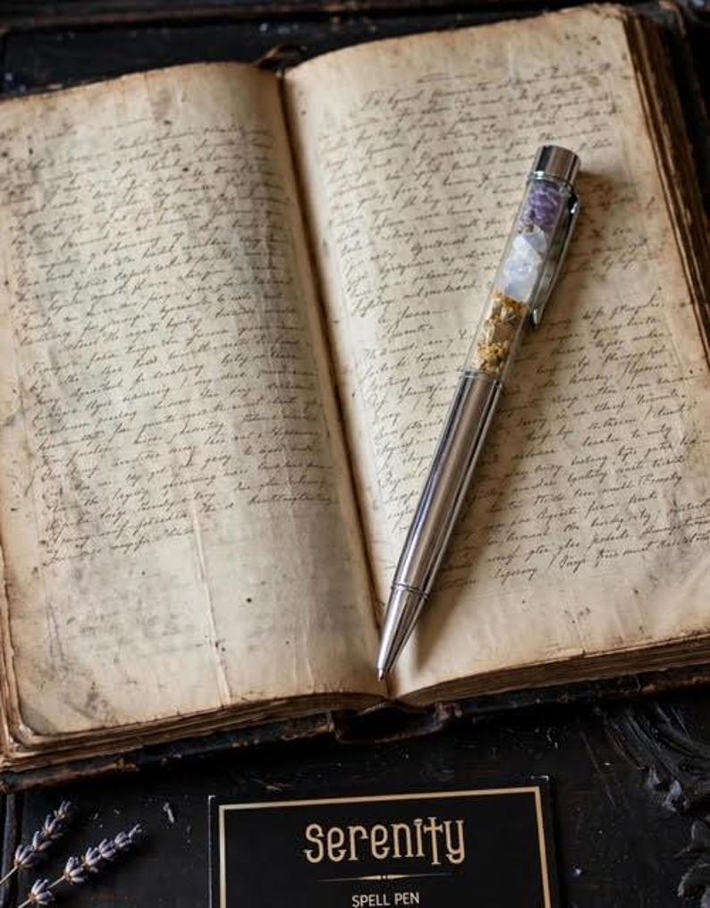 May include: A silver-toned ballpoint pen with a clear upper barrel containing small purple, white, and gold-coloured decorative fragments, resting on an open antique book with handwritten cursive text on aged, yellowed paper pages.