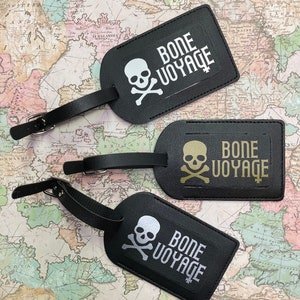 May include: Three black leather luggage tags with a skull and crossbones design and the text "Bone Voyage". The tags have different coloured skulls: white, gold, and silver.