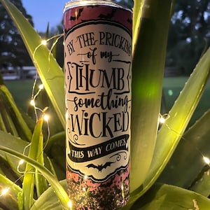 May include: A pink and black tumbler with a white sticker that says "By the pricking of my thumb, something wicked this way comes".