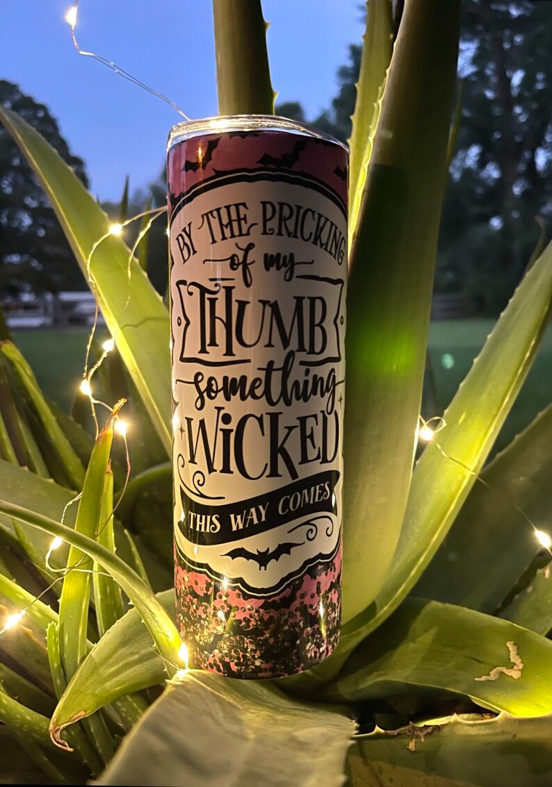 May include: A pink and black tumbler with a white sticker that says "By the pricking of my thumb, something wicked this way comes".