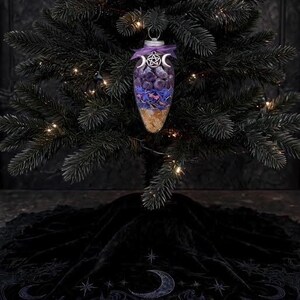 May include: A clear, teardrop-shaped glass ornament filled with layers of purple stones, blue flower petals, and yellow crystals. The top is adorned with a purple ribbon and a silver-toned metal charm featuring a pentagram and crescent moons.