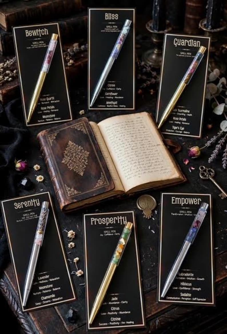 May include: Five spell pens with clear barrels filled with crystals and botanicals, displayed on black cards labelled Bewitch, Bliss, Guardian, Serenity, Prosperity, and Empower, surrounding an open, leather-bound journal with gold embossed details.