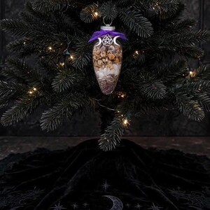May include: A clear, teardrop-shaped glass ornament filled with dried flowers and white crystals. The ornament is topped with a purple ribbon and a silver-toned metal charm featuring a central pentagram flanked by two crescent moons.