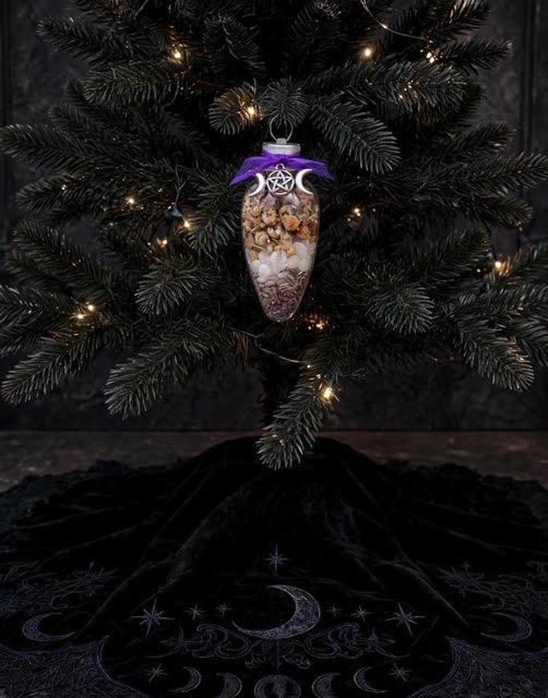 May include: A clear, teardrop-shaped glass ornament filled with dried flowers and white crystals. The ornament is topped with a purple ribbon and a silver-toned metal charm featuring a central pentagram flanked by two crescent moons.