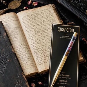 May include: A gold-toned pen with a clear barrel filled with black tourmaline, rose petals, and tiger's eye, resting on a black card labelled Guardian Spell Pen. The card lists the contents and their properties next to an open, aged, handwritten book.