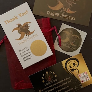 May include: A dark red organza drawstring pouch, a white scratch-off card with gold text and a winged unicorn logo, a gold rectangular card with a winged unicorn, a circular sticker, and a black business card with gold accents and a QR code.