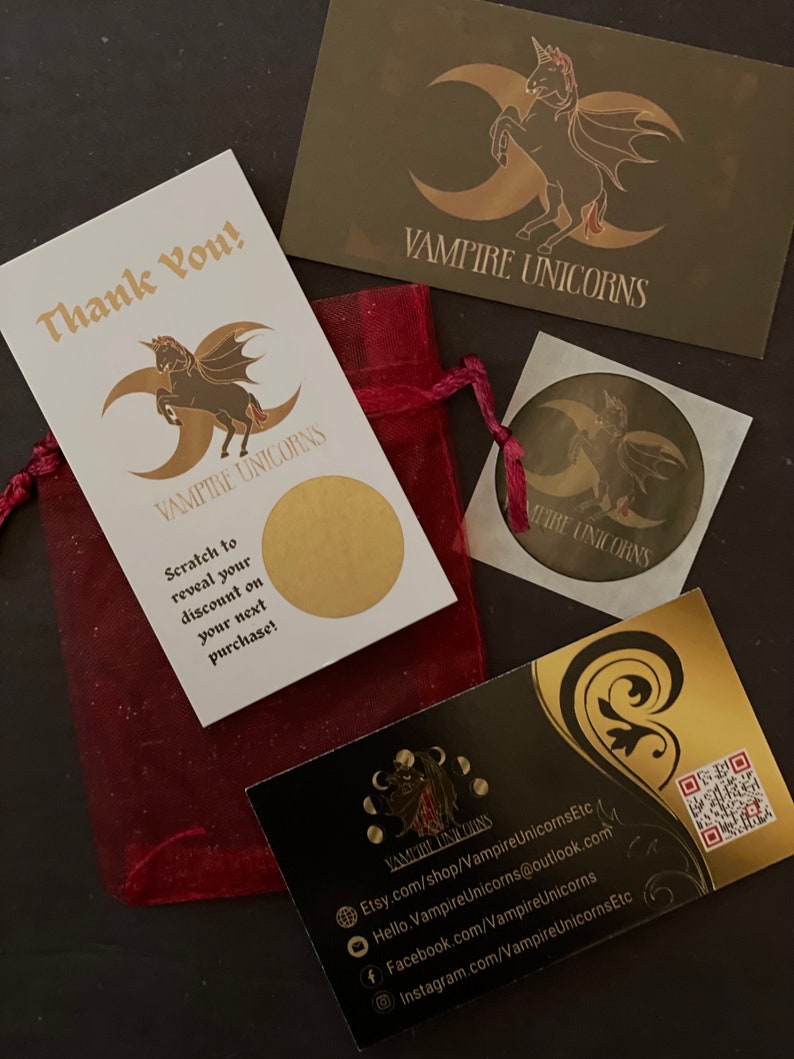 May include: A dark red organza drawstring pouch, a white scratch-off card with gold text and a winged unicorn logo, a gold rectangular card with a winged unicorn, a circular sticker, and a black business card with gold accents and a QR code.