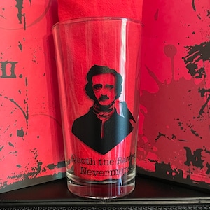 May include: A clear glass with a black silhouette of Edgar Allan Poe and a raven. The glass has the text "Quoth the Raven Nevermore" printed on the bottom.