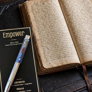 May include: A silver-toned spell pen with a clear barrel containing small crystals, resting on a black product card labelled Empower, Labradorite, Hibiscus, and Sodalite, positioned next to an open antique book with handwritten cursive text on aged pages.