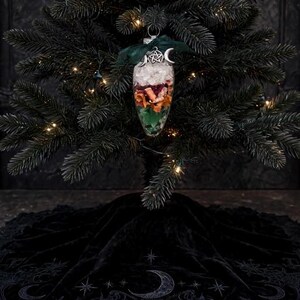 May include: A clear, teardrop-shaped glass ornament filled with layers of white crystals, dried orange slices, and green moss. It is topped with a dark green ribbon and a silver pentacle charm with crescent moons, hanging on a dark green tree.
