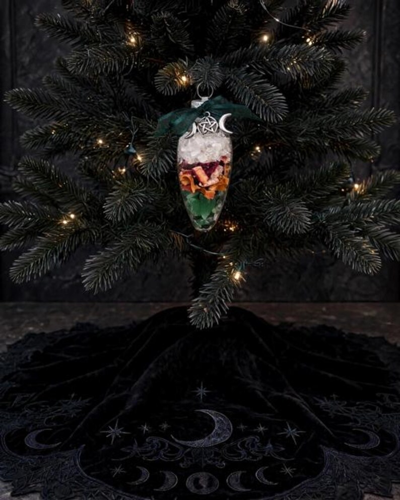 May include: A clear, teardrop-shaped glass ornament filled with layers of white crystals, dried orange slices, and green moss. It is topped with a dark green ribbon and a silver pentacle charm with crescent moons, hanging on a dark green tree.
