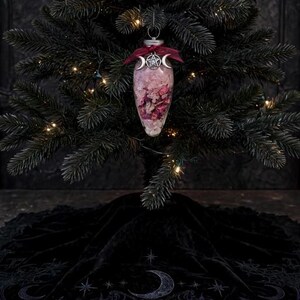 May include: A clear, teardrop-shaped glass ornament filled with pink flower petals and crystals, topped with a dark red ribbon and a silver-toned metal charm featuring a central pentagram flanked by two crescent moons, hanging on a tree.