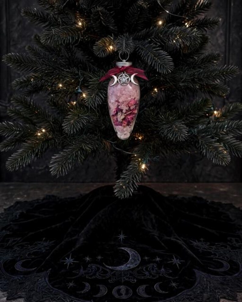 May include: A clear, teardrop-shaped glass ornament filled with pink flower petals and crystals, topped with a dark red ribbon and a silver-toned metal charm featuring a central pentagram flanked by two crescent moons, hanging on a tree.