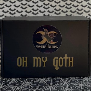 Op de afbeelding: Zwarte doos met een gouden ontwerp. De doos heeft een ronde sticker met een vampier eenhoorn afbeelding en de tekst "VAMPIRE UNICORNS". De woorden "OH MY GOTH" staan eronder in goudfolie.