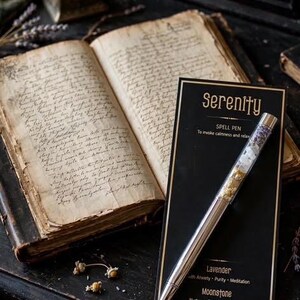 May include: A silver ballpoint pen with a clear barrel filled with dried lavender, moonstone, and chamomile, displayed on a black card labelled Serenity Spell Pen with text listing Lavender, Moonstone, and Chamomile properties for calm and relaxation.