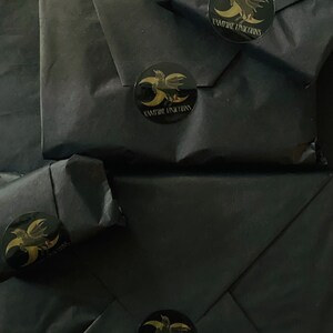May include: Four packages wrapped in matte black paper, each sealed with a circular black sticker featuring a gold graphic of a winged unicorn and a crescent moon, with the text VAMPIRE UNICORNS printed in gold lettering below the graphic.