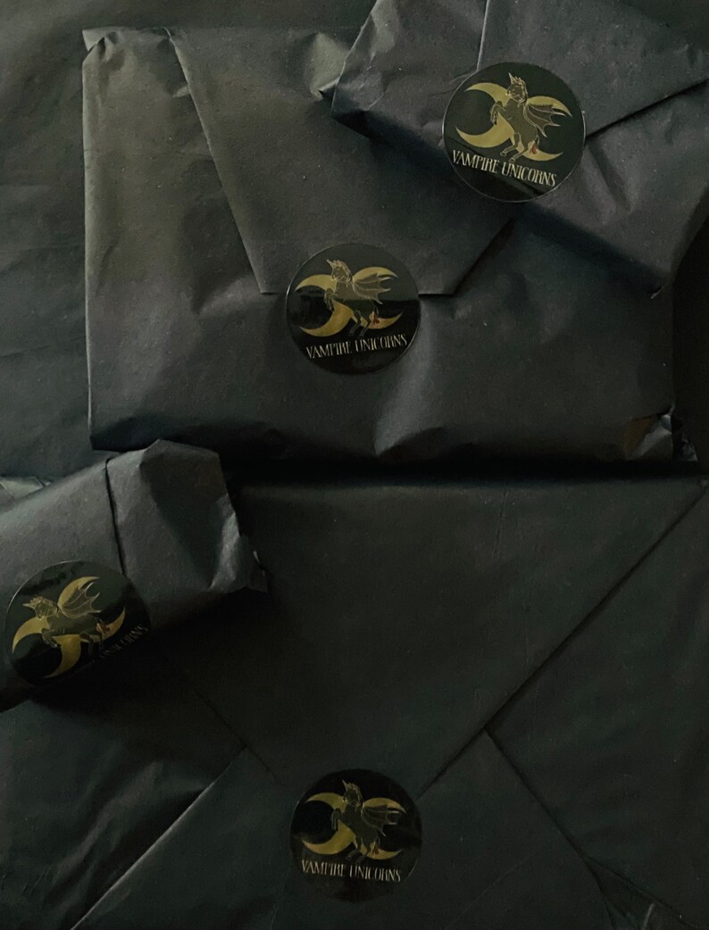 May include: Four packages wrapped in matte black paper, each sealed with a circular black sticker featuring a gold graphic of a winged unicorn and a crescent moon, with the text VAMPIRE UNICORNS printed in gold lettering below the graphic.
