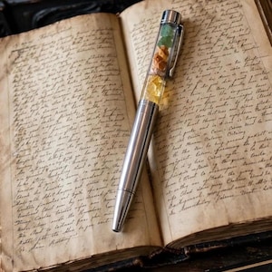 May include: A silver-toned ballpoint pen with a clear upper barrel containing small, loose, multi-coloured gemstone fragments in green, orange, and yellow. The pen features a metallic clip and rests diagonally across an open, aged, handwritten book.