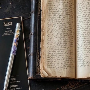 May include: A silver-toned pen with a clear barrel filled with small citrine, cornflower, and amethyst crystals, resting on a black card labelled BIISS SPELL PEN, next to an open antique book with aged, yellowed pages covered in handwritten cursive text.