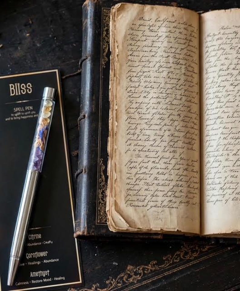 May include: A silver-toned pen with a clear barrel filled with small citrine, cornflower, and amethyst crystals, resting on a black card labelled BIISS SPELL PEN, next to an open antique book with aged, yellowed pages covered in handwritten cursive text.
