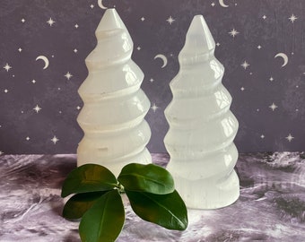 May include: Two white selenite crystal towers with a spiral design, resembling trees, on a gray and white marbled surface with a green leaf sprig in front.