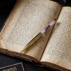May include: A gold-toned ballpoint pen with a clear barrel filled with small pink and white floral petals, resting diagonally across the open pages of an antique book featuring handwritten cursive text in dark ink on aged, yellowed paper.