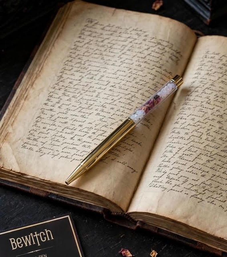 May include: A gold-toned ballpoint pen with a clear barrel filled with small pink and white floral petals, resting diagonally across the open pages of an antique book featuring handwritten cursive text in dark ink on aged, yellowed paper.