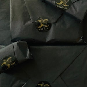 May include: Four packages wrapped in matte black paper, each sealed with a circular black sticker featuring a gold graphic of a winged unicorn and a crescent moon, with the text VAMPIRE UNICORNS printed in gold lettering below the graphic.