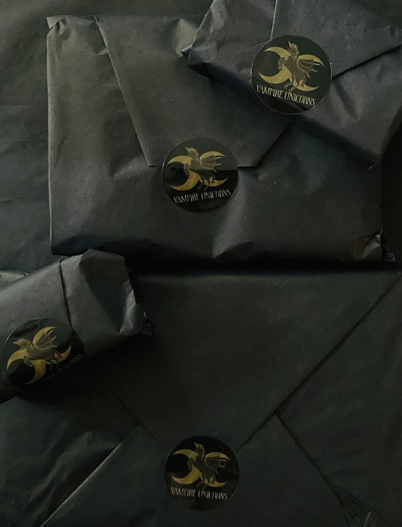 May include: Four packages wrapped in matte black paper, each sealed with a circular black sticker featuring a gold graphic of a winged unicorn and a crescent moon, with the text VAMPIRE UNICORNS printed in gold lettering below the graphic.