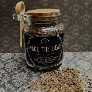 May include: A glass jar with a cork lid and a wooden spoon, filled with a dark, granular substance. The jar has a black label that reads "WAKE THE DEAD" and lists ingredients. A small pile of the substance sits next to the jar. The background features a skull pattern.